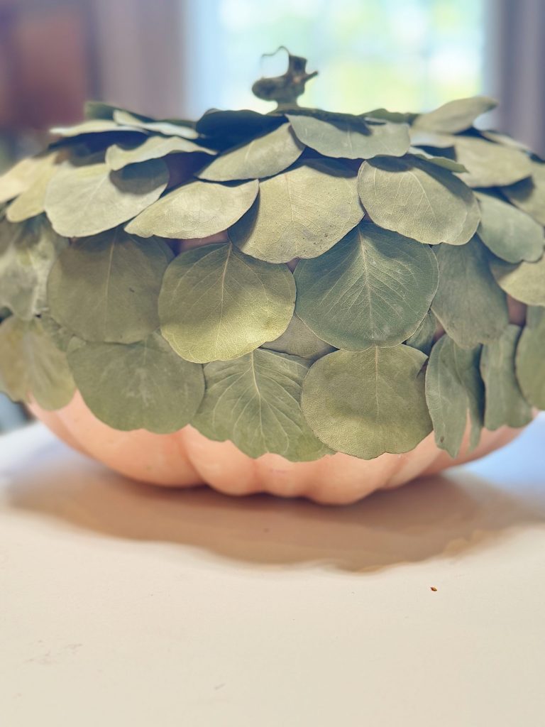 A pumpkin covered with green leaves, arranged in a layered pattern, sits on a light-colored surface.