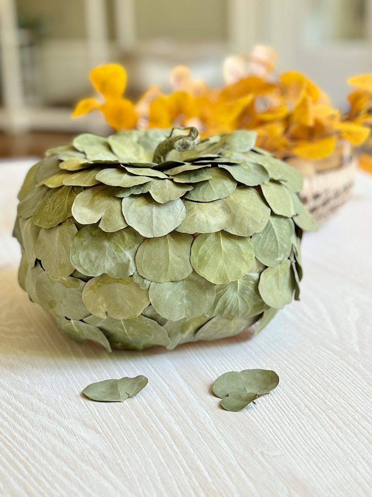 A pumpkin decorated with layered green leaves sits on a white surface. A few leaves are scattered nearby. Background features blurred yellow foliage.