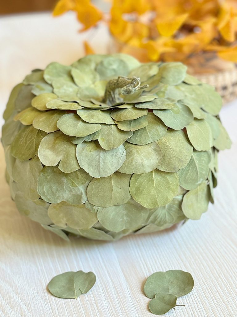 A decorative pumpkin covered with green dried leaves is placed on a light wooden surface, with a cluster of yellow leaves in the background.