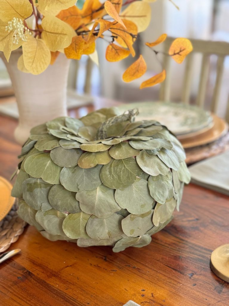 A decorative pumpkin made of layered green leaves sits on a wooden table, accompanied by plates and a vase with yellow leaves.