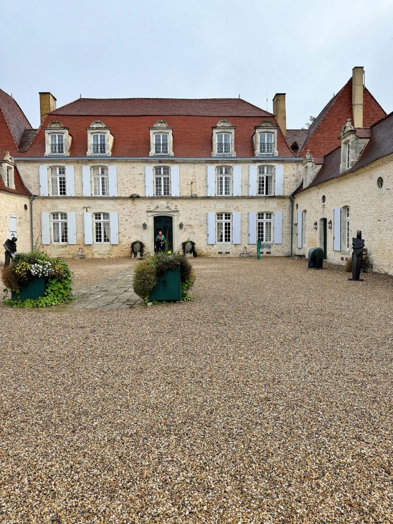 A large historic building with white walls, red roof, and multiple windows. There is a courtyard with gravel, greenery, and statues in front of the building.