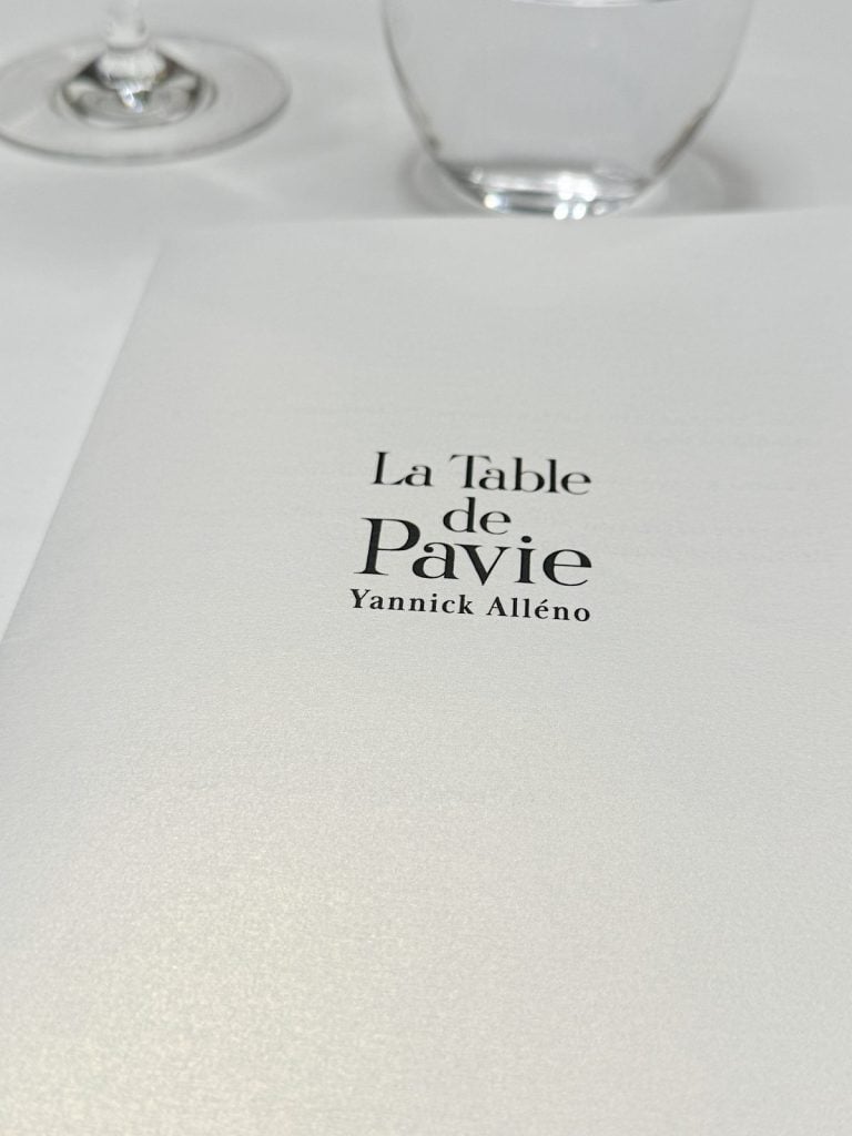 Close-up of a menu from "La Table de Pavie" by Yannick Alléno, with blurred wine glass and water glass in the background.