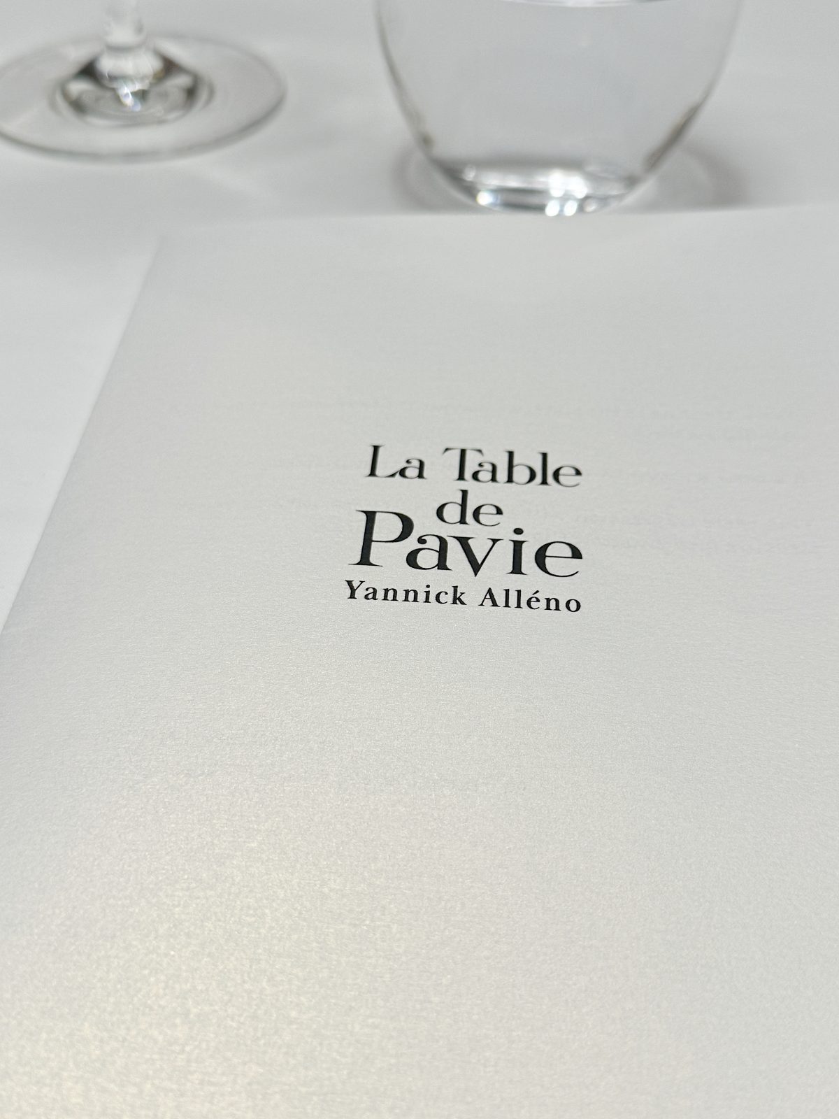 Close-up of a menu from "La Table de Pavie" by Yannick All&eacute;no, with blurred wine glass and water glass in the background.
