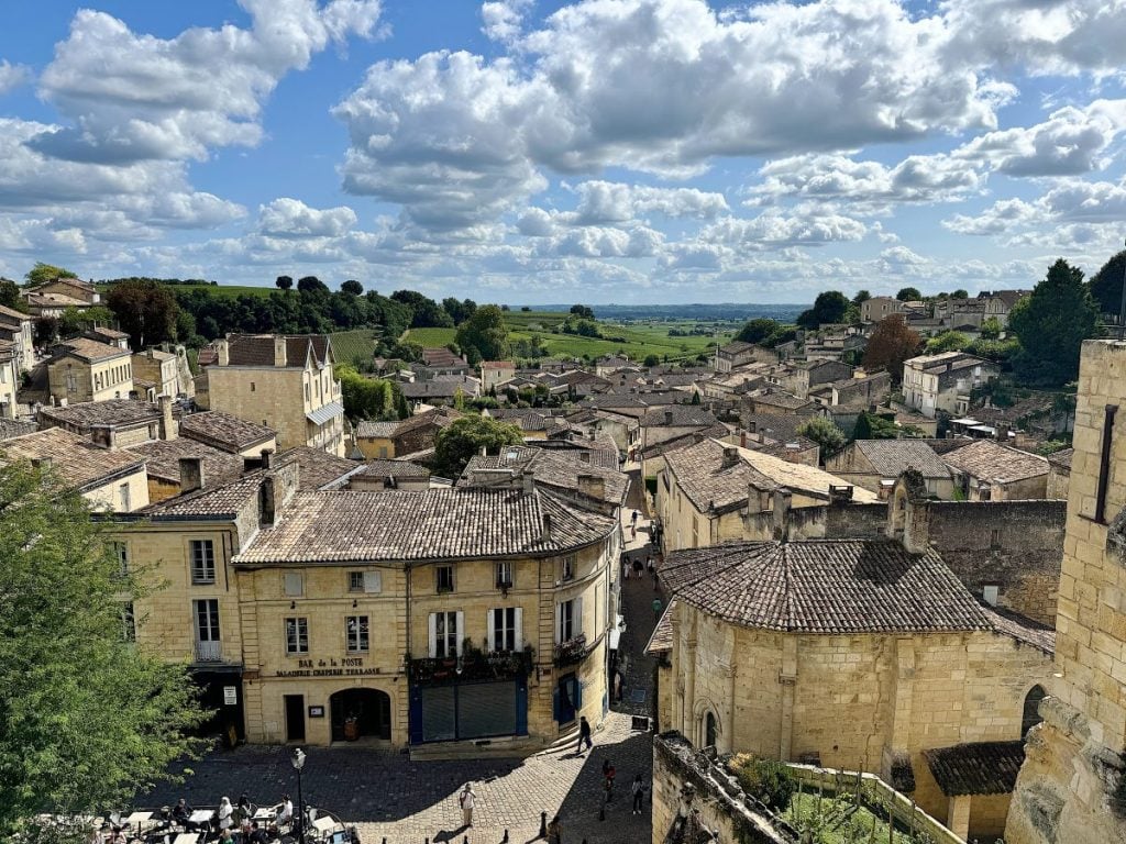 A panoramic view of a historic town with beige stone buildings and tiled roofs. Rolling green fields extend into the distance under a partly cloudy sky. People walk along a cobblestone street below.