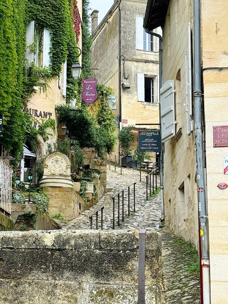 Narrow cobblestone street in a historic town lined with old buildings, greenery, and restaurant signs. Stone steps with handrails lead uphill.