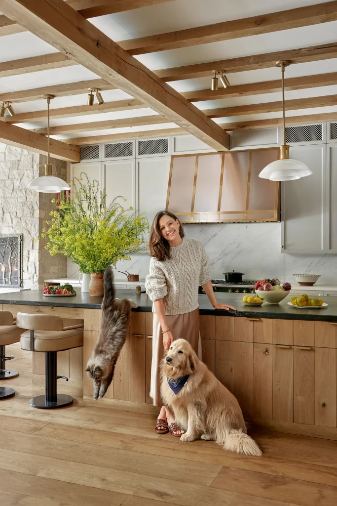 A woman stands in a kitchen, smiling and petting a golden retriever. A cat jumps down from a kitchen island. The kitchen features wooden beams, bar stools, and various items on the counter.