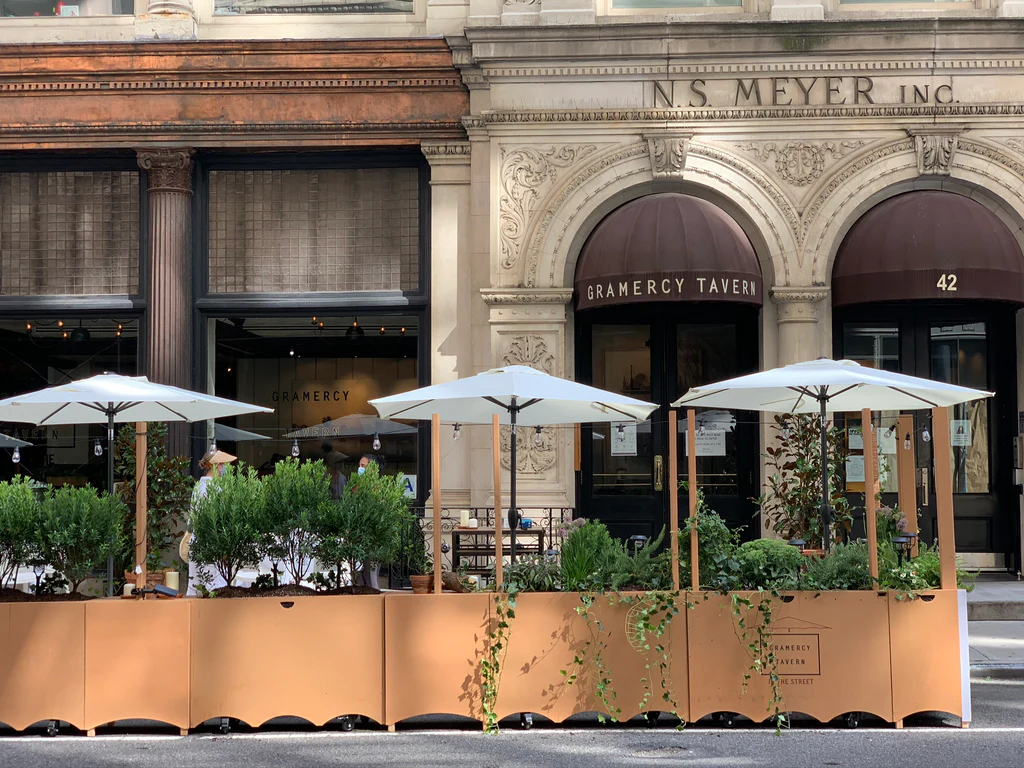 Outdoor dining area of Gramercy Tavern with white umbrellas and potted plants in front of a historic building facade.