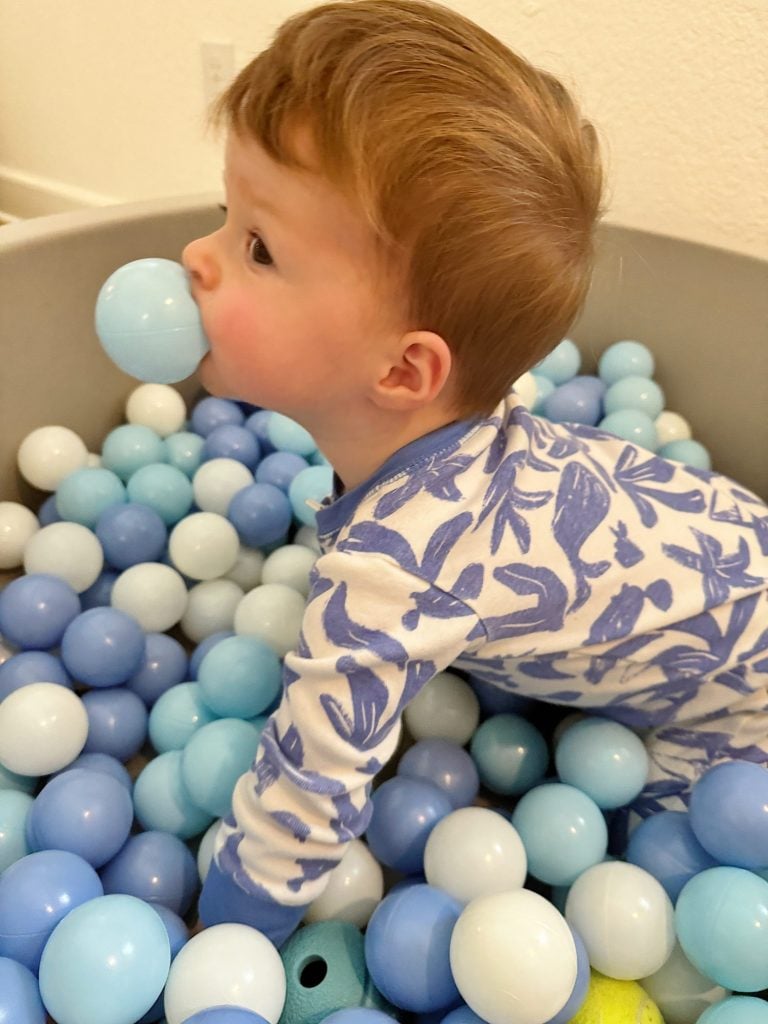 A young child in a blue and white outfit is playing in a ball pit filled with various shades of blue and white balls, holding a blue ball in their mouth.