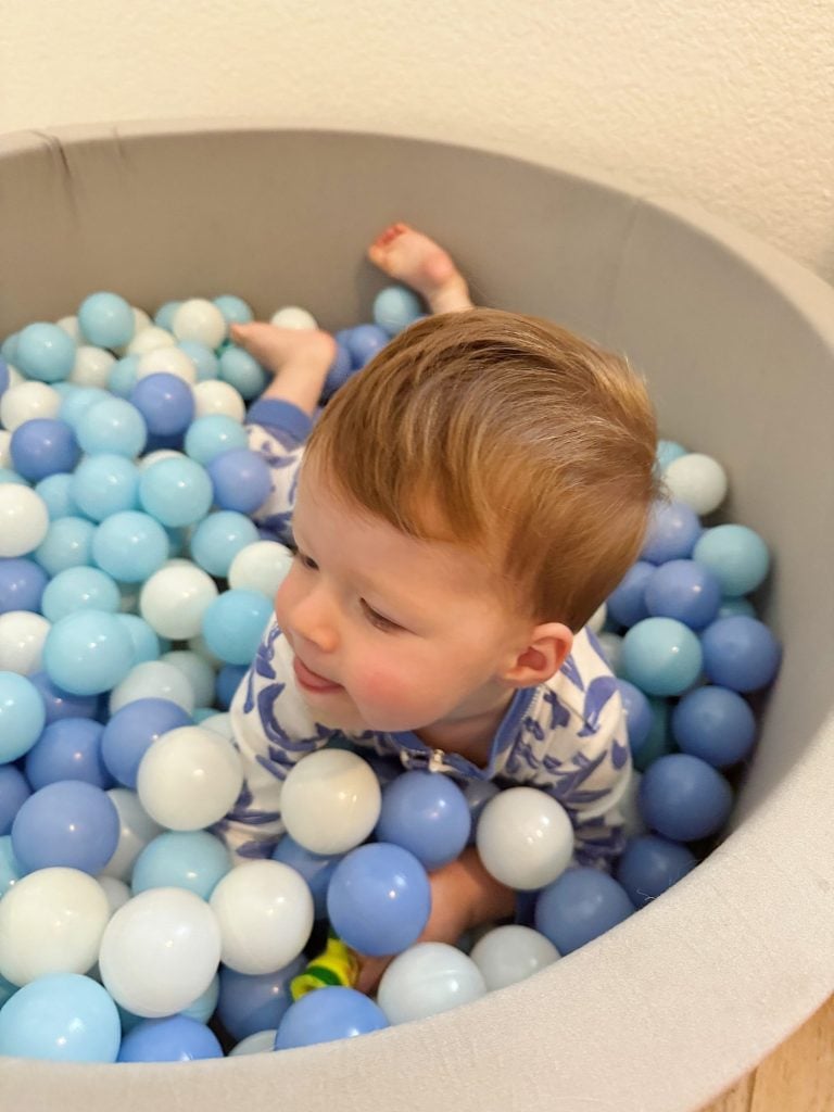 A toddler in pajamas sits in a ball pit filled with blue, white, and light blue balls, looking to the side.