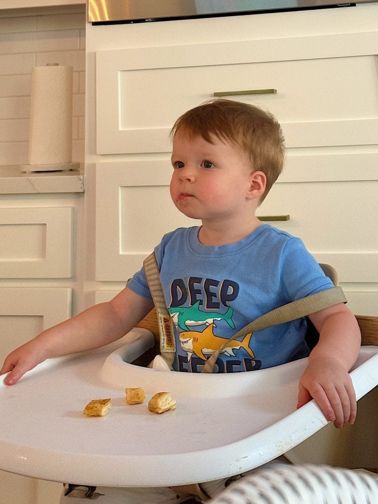 A young child sits in a high chair wearing a blue shirt with "DEEP SLEEPER" text. Pieces of food are on the tray in front of them.