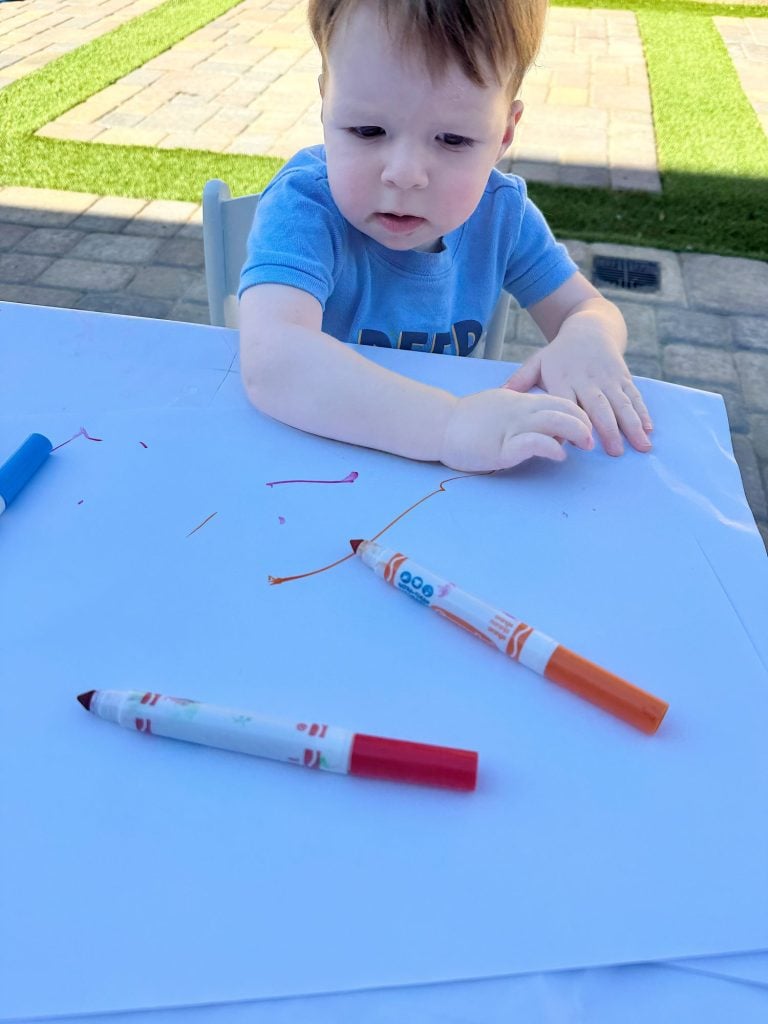 A child in a blue shirt draws on a large sheet of white paper with red and orange markers under outdoor sunlight.