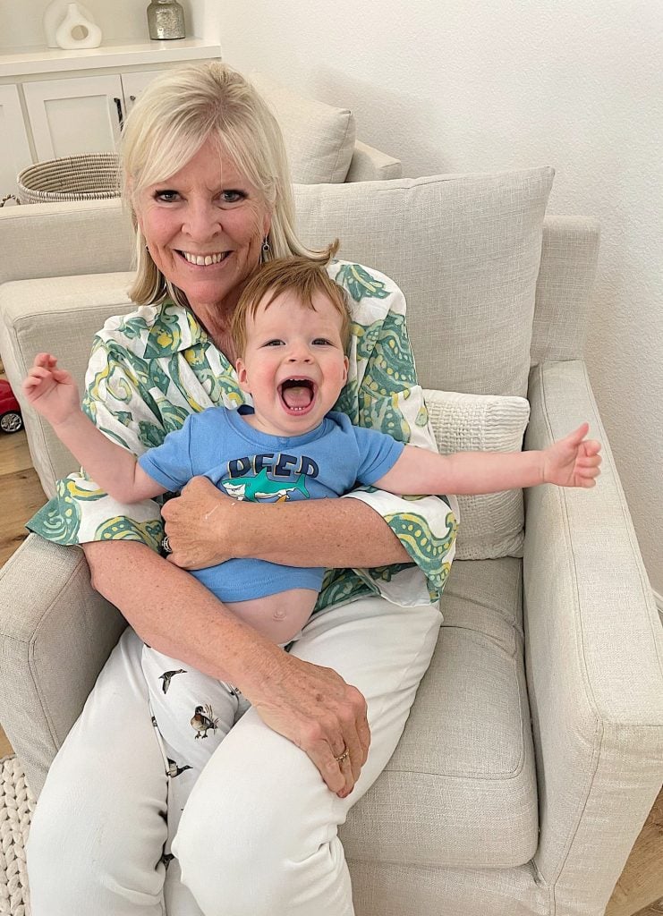 A woman sitting on a chair and smiling, holds a laughing toddler on her lap. Both appear happy and are dressed in casual clothes.