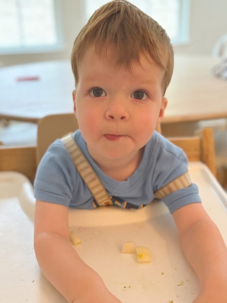 A child in a blue shirt sits in a high chair, looking at the camera with pieces of food on the tray in front of him.