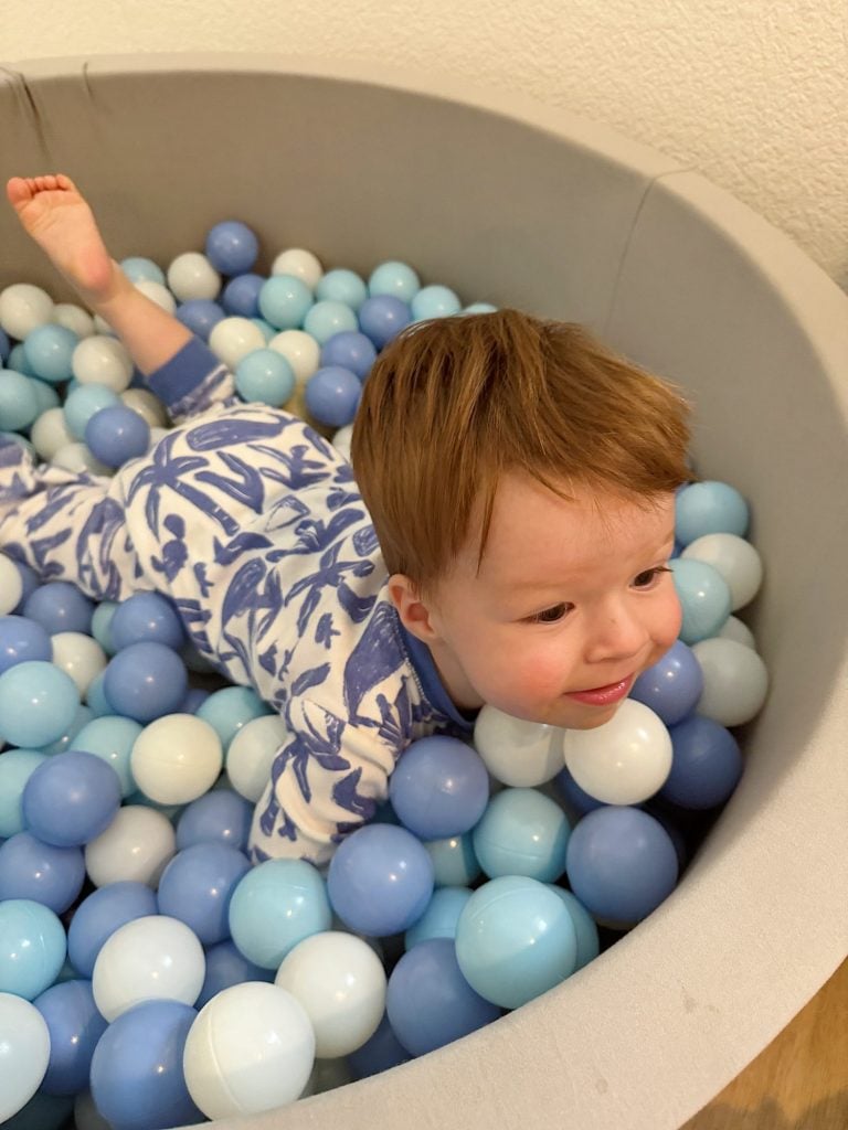 A toddler in patterned pajamas lies in a ball pit filled with blue and white plastic balls, smiling contentedly.