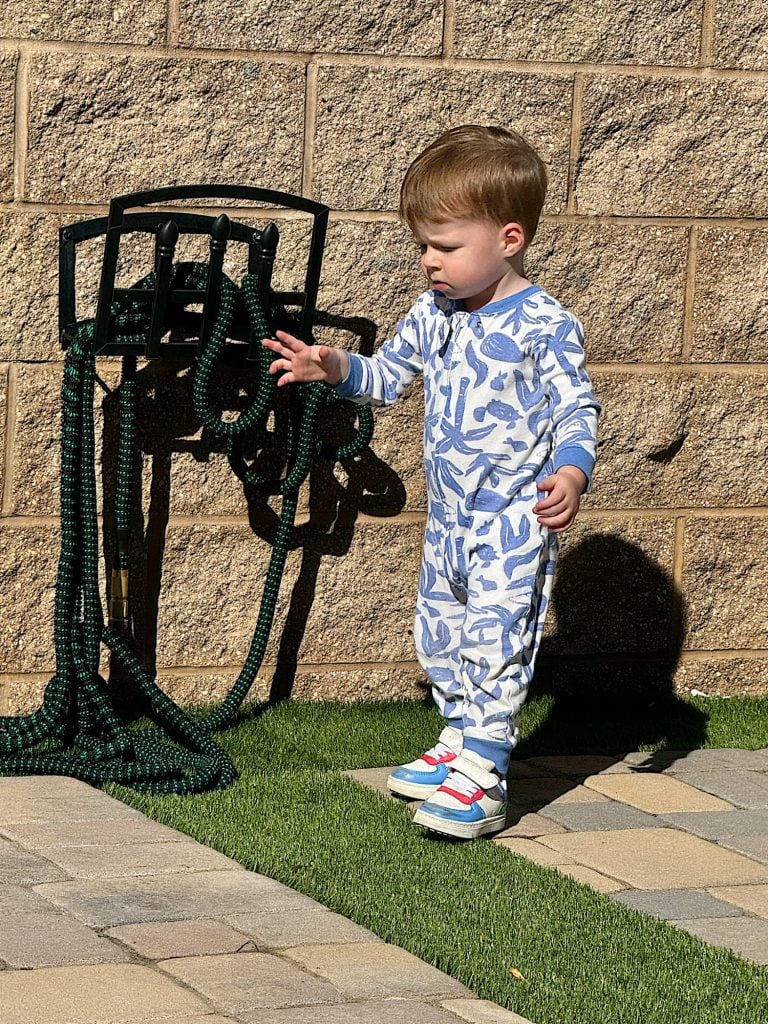 A young child in pajamas stands on artificial grass near a wall and holds a garden hose. The child appears to be reaching out with one hand.