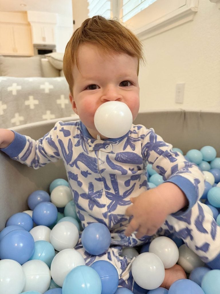 A young child in blue and white pajamas is sitting in a ball pit, holding a ball in their mouth, surrounded by various colored balls.