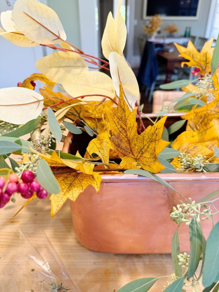 A copper container holds an arrangement of yellow maple leaves, white and green foliage, and small clusters of pink berries on a wooden table.