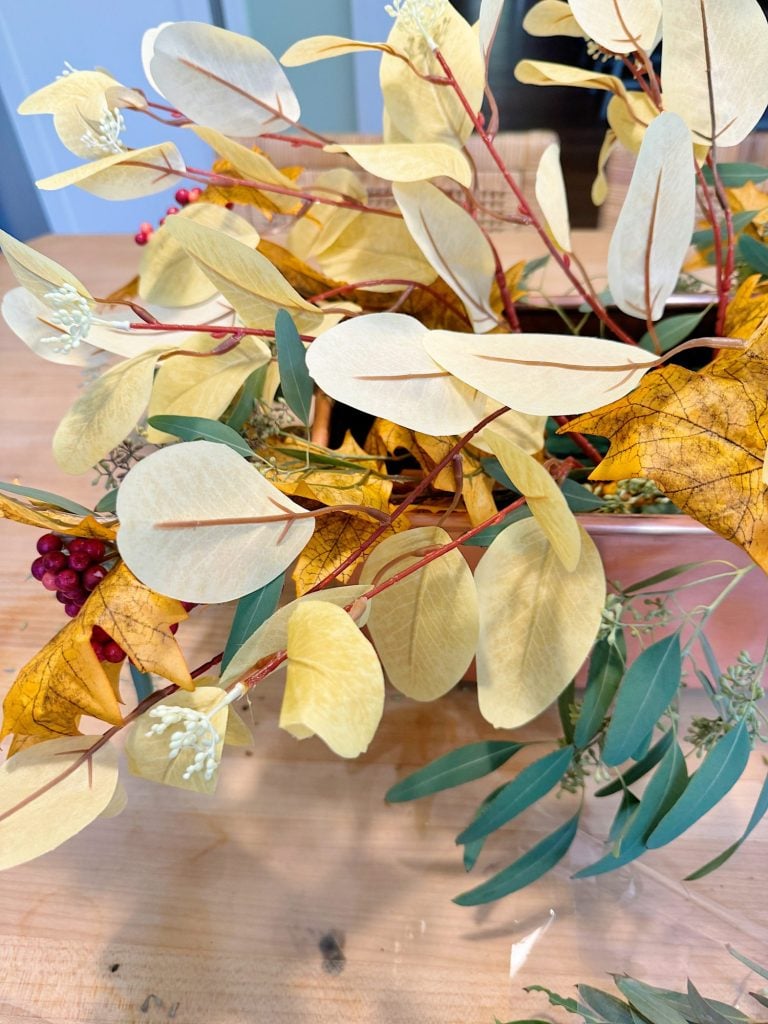 A bouquet of dried yellow leaves with some green foliage and red berries arranged in a container on a wooden surface.