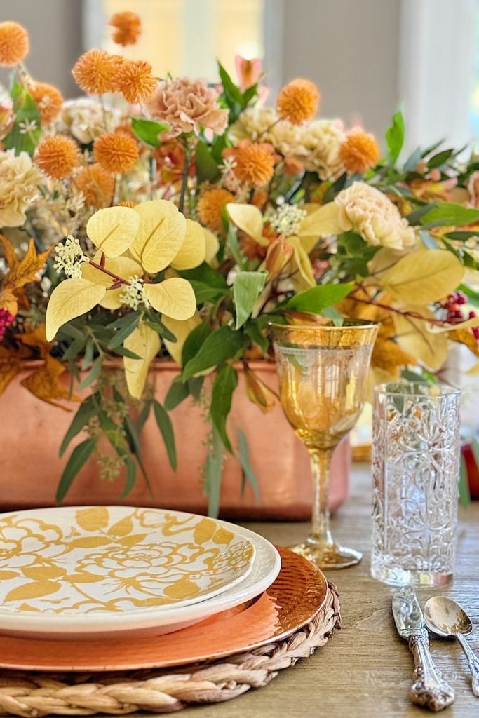 A dining table set with a yellow and white plate, a glass goblet, a clear drinking glass, cutlery, and a floral arrangement with yellow and orange flowers.