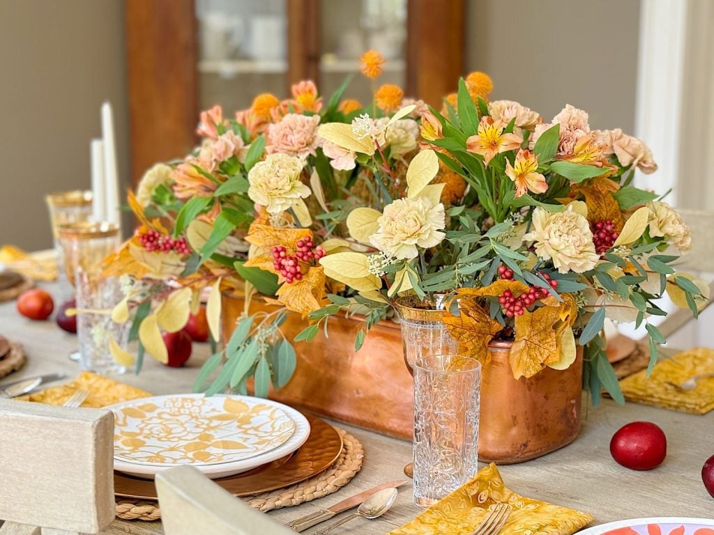 A dining table set with patterned plates, crystal glasses, and yellow napkins, featuring a large copper centerpiece containing an arrangement of yellow, orange, and pink flowers with greenery.