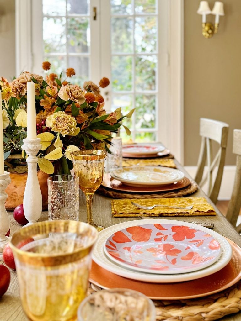 A dining table set for four with floral plates, amber glassware, and a centerpiece of flowers. The scene includes light streaming through a large window in the background.