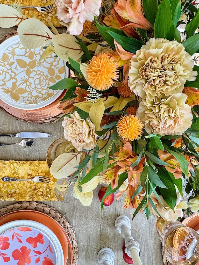 A dining table is set with floral-patterned plates, yellow napkins, and a vibrant centerpiece featuring a mix of large pastel and orange flowers with green leaves.