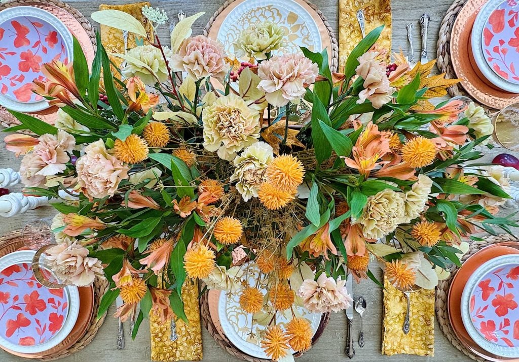 A table set for a meal, featuring a centerpiece with pink, orange, and yellow flowers, surrounded by plates, silverware, and floral-themed napkins.