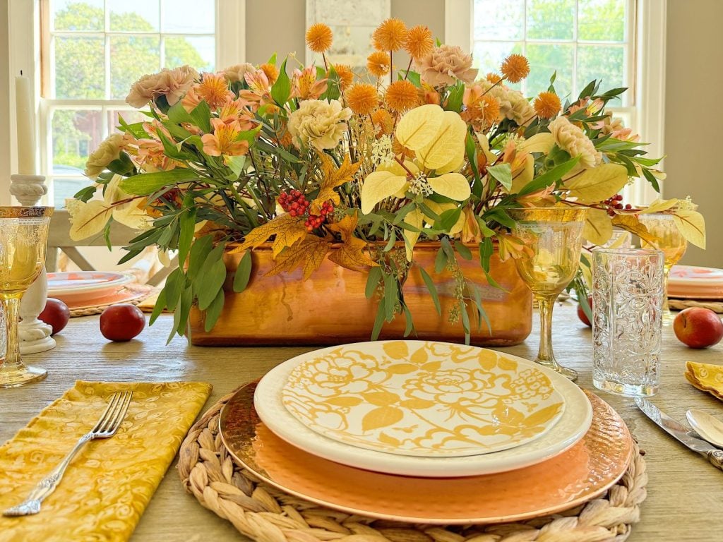 A table setting with a floral centerpiece in a copper container, yellow and white decorative plates on wicker chargers, yellow napkins, and glasses is bathed in natural light.