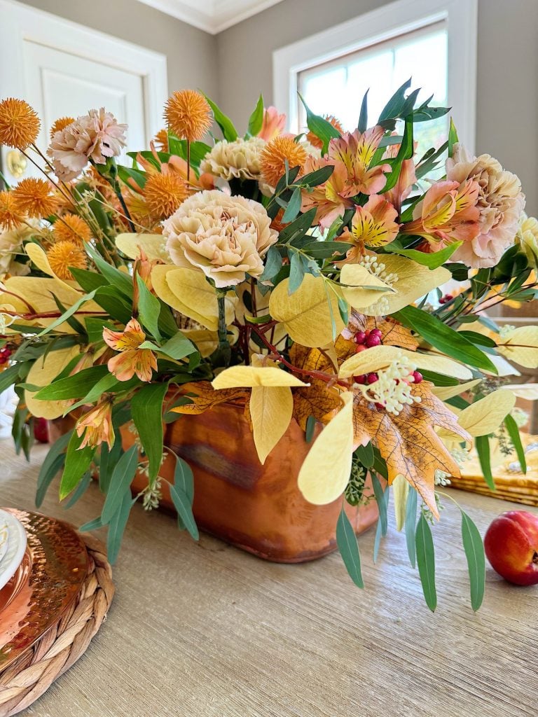 A fall-themed floral arrangement in a copper vase sits on a wooden table. The bouquet features orange, yellow, and white flowers with greenery and autumn leaves.