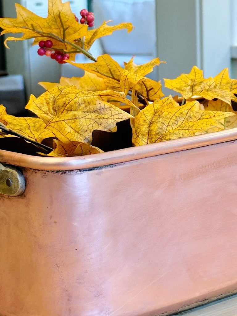 A copper planter is filled with vibrant yellow autumn leaves and small red berries, displayed indoors.