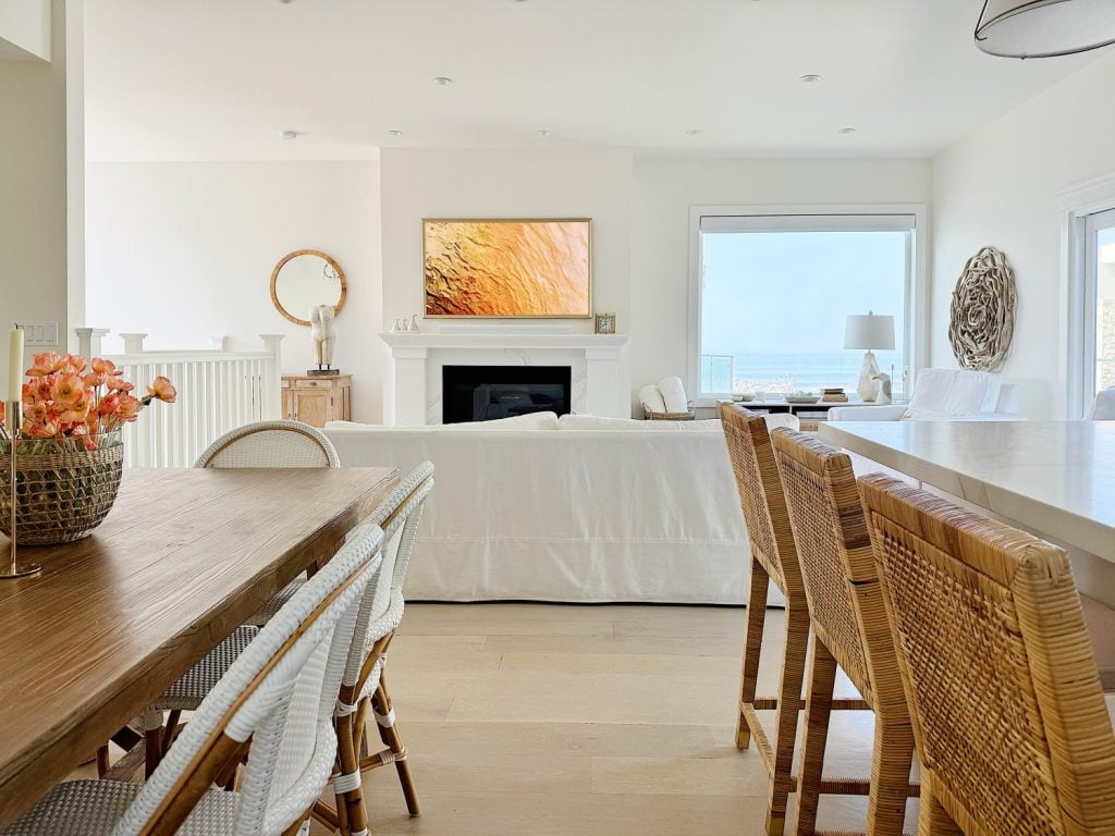 A spacious, bright living area with white walls and furniture, a wooden dining table with chairs, wicker bar stools, a fireplace, and a wall-mounted TV. Large window offers a view of the ocean.