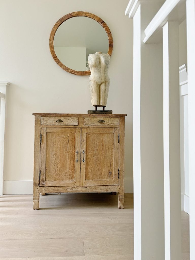 A wooden cabinet with two drawers and double doors is topped with a mannequin torso sculpture. Above hangs a round mirror with a wooden frame. White stair balusters are visible on the right.