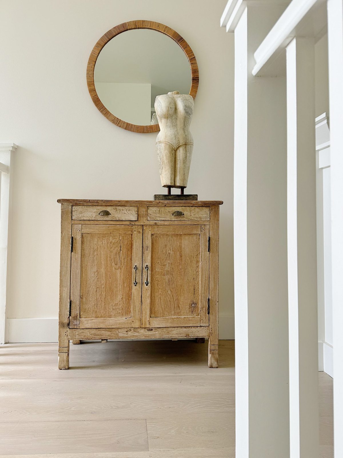 A wooden cabinet with two drawers and double doors is topped with a mannequin torso sculpture. Above hangs a round mirror with a wooden frame. White stair balusters are visible on the right.