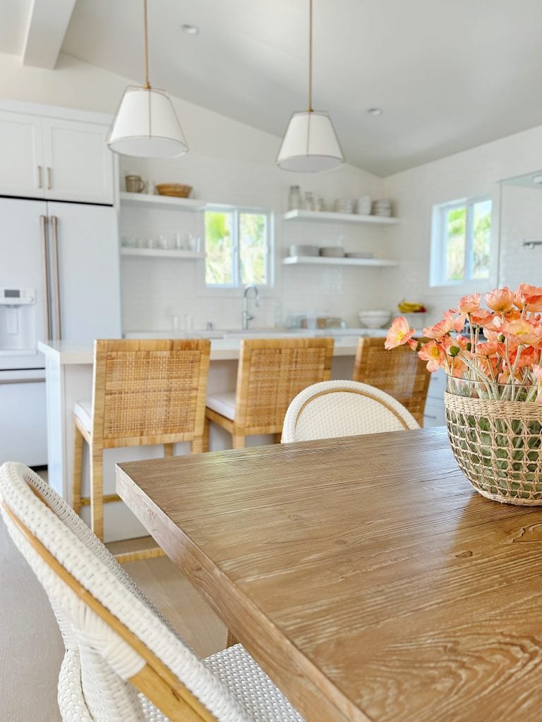 A bright kitchen with wicker chairs, white cabinetry, and a wooden dining table with a floral centerpiece. Two pendant lights hang above the island.