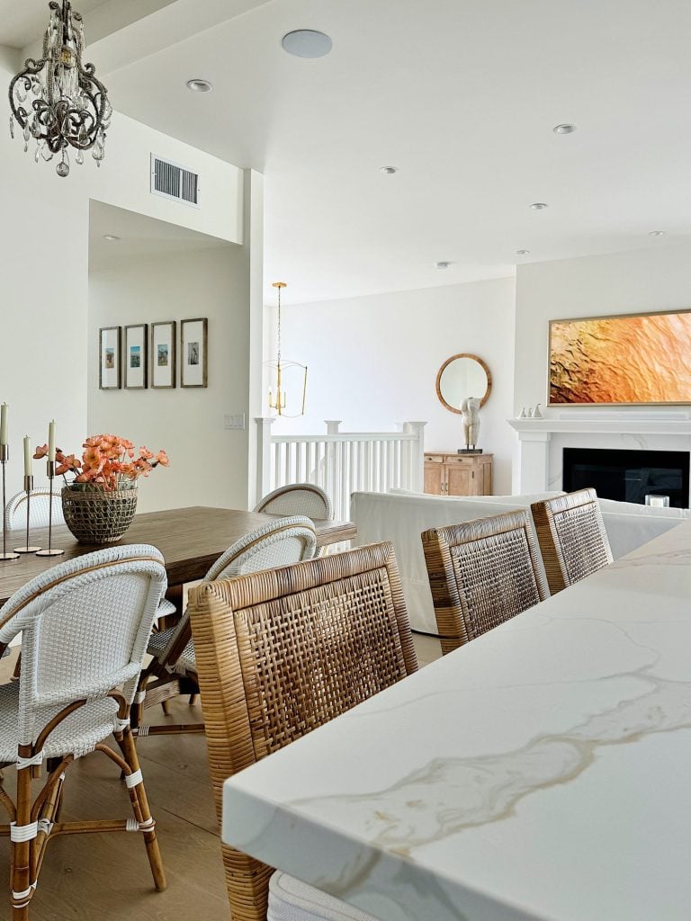 A modern open-plan dining and living area with a marble countertop, wicker and cushioned chairs, a wooden dining table, framed photos on the wall, a chandelier, and a television over a fireplace.