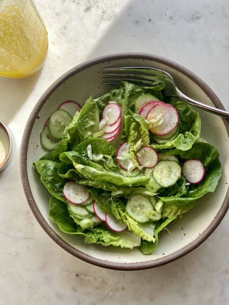 A bowl of green salad with lettuce, sliced cucumbers, and radishes, accompanied by a fork, sits on a white surface.