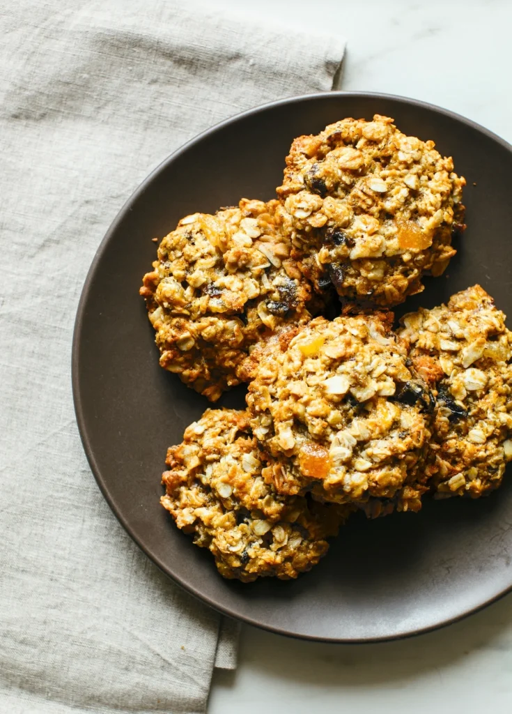 A dark plate holding four homemade oatmeal cookies with raisins and nuts rests on a beige cloth.