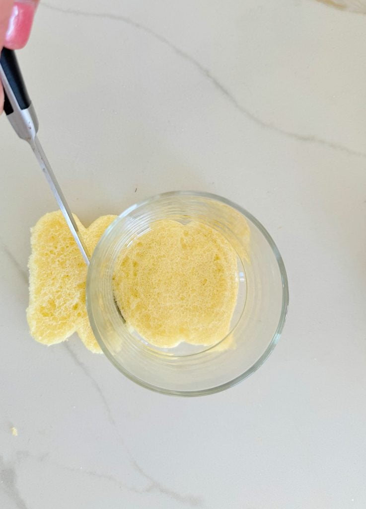 A slice of yellow sponge cake is being cut into a circular shape using a drinking glass and a knife on a light countertop.