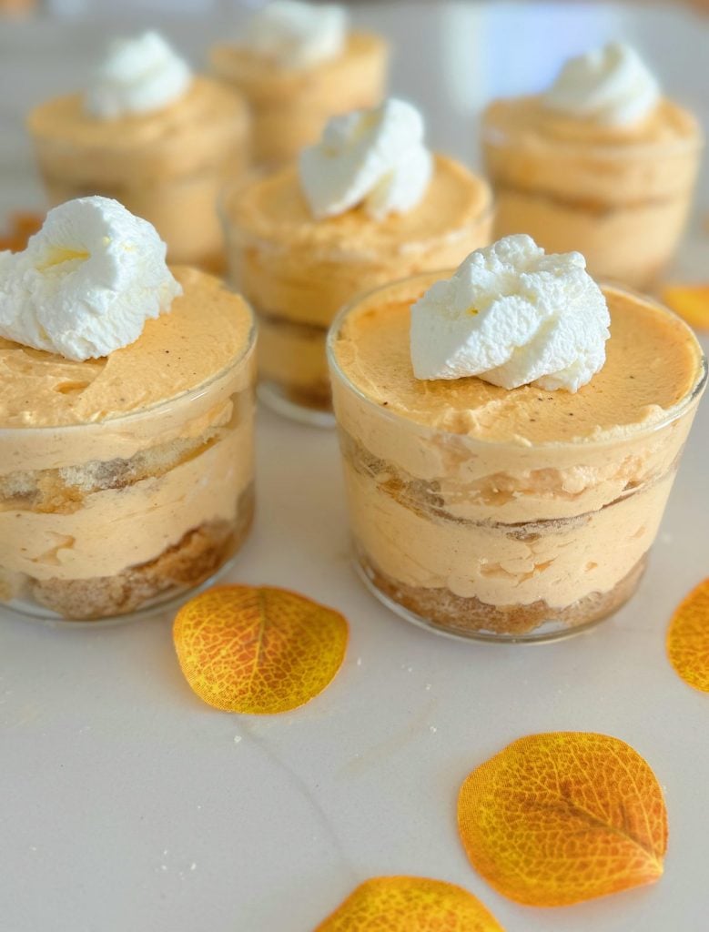 Five small dessert cups filled with layered pumpkin mousse and topped with whipped cream, surrounded by yellow autumn leaves on a white surface.