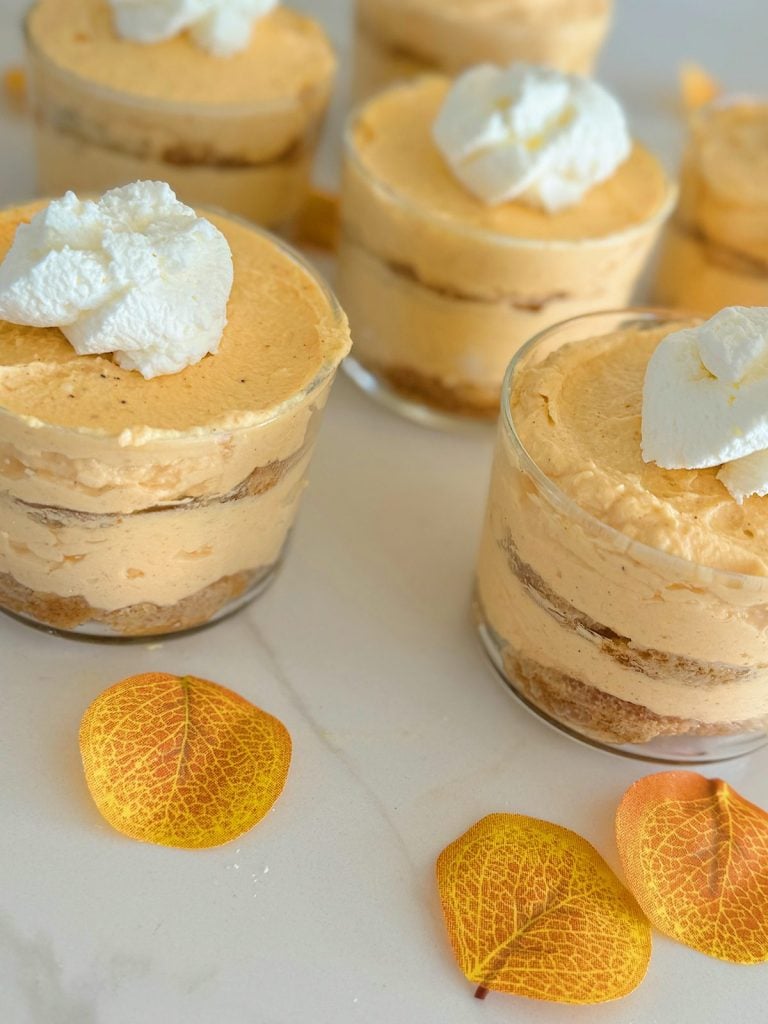 Four glass cups filled with layered pumpkin mousse and topped with whipped cream, placed on a white surface with three autumn leaves scattered nearby.