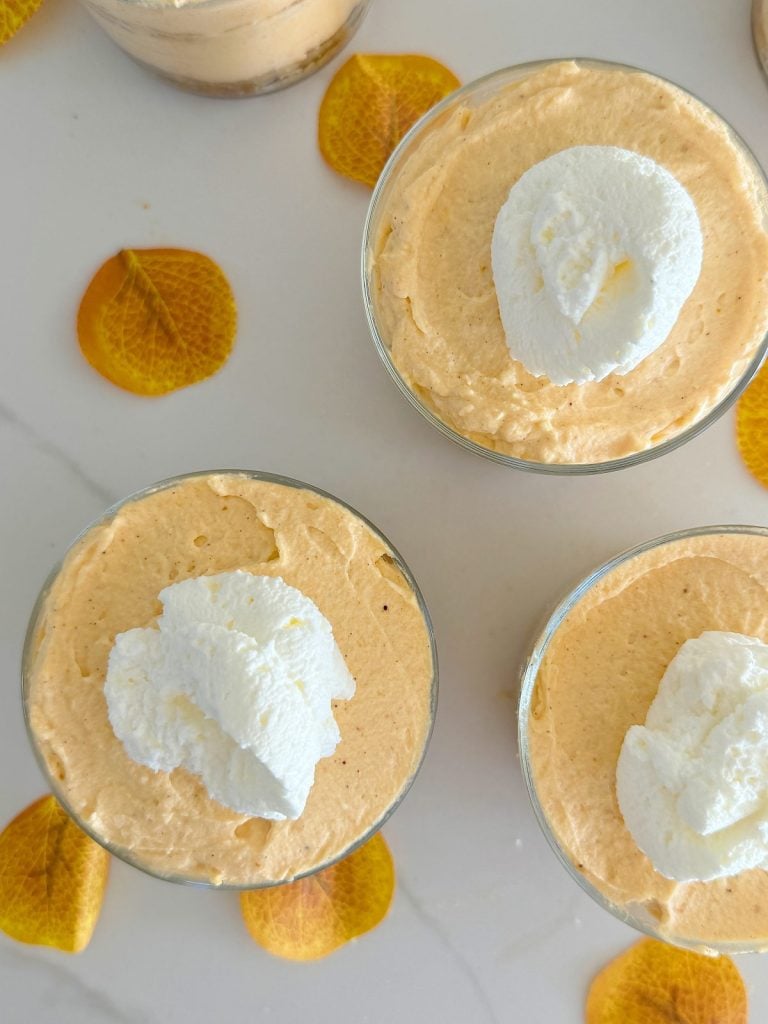 Three glasses of pumpkin mousse topped with dollops of whipped cream are surrounded by scattered yellow leaves on a white surface.