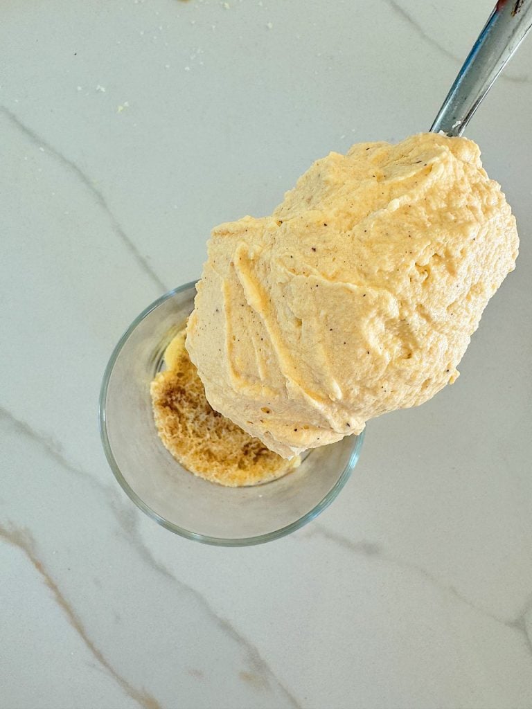 A large scoop of thick, beige batter is being placed into a glass bowl containing a small amount of brown powder on a light-colored countertop.