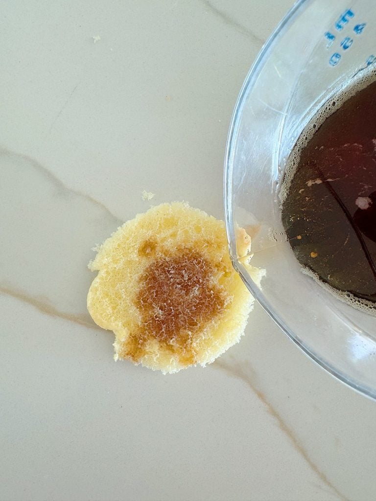 A close-up of a spilled beverage forming a circular stain on a light-colored surface next to a glass measuring cup.