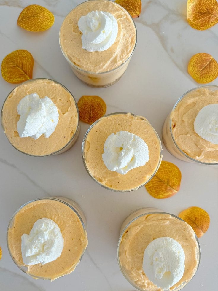 Six cups of pumpkin mousse topped with whipped cream are arranged on a white marble surface, surrounded by decorative fall leaves.