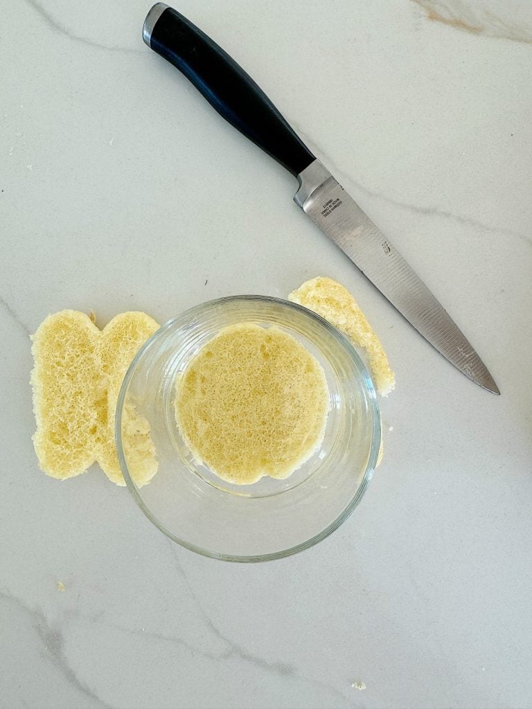 A bread slice with a circular piece cut out is on a marble countertop; a knife with a black handle is placed next to it, and a glass bowl is positioned over the hole in the bread.