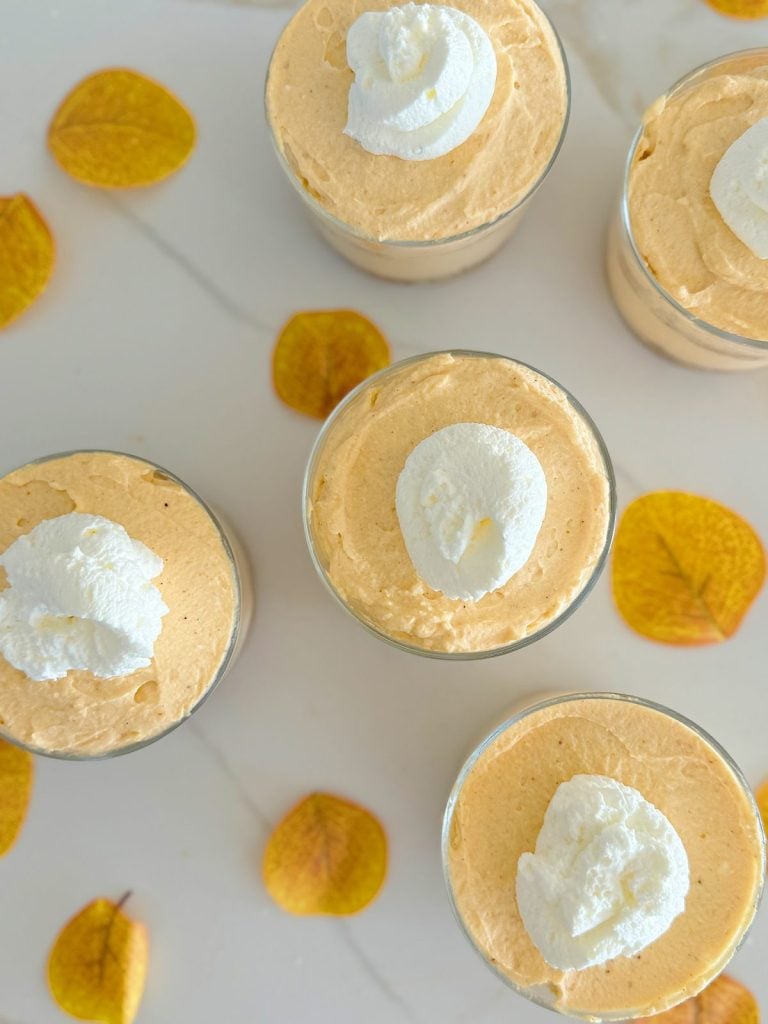 Five glasses of pumpkin mousse topped with whipped cream are arranged on a white surface, surrounded by scattered autumn leaves.