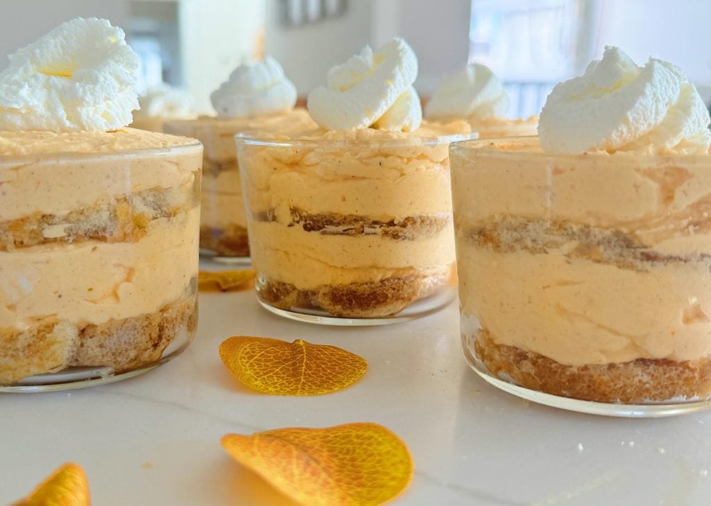 Individual dessert cups filled with layers of creamy pumpkin mousse, whipped cream, and crumbled cookies, garnished with dollops of whipped cream. Dried orange leaves scattered on the table.