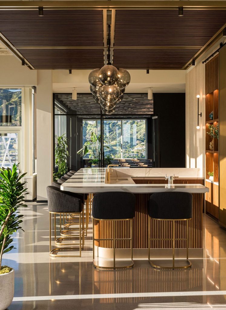 Modern kitchen with a central island featuring bar stools, pendant lights, and a view of mountains through large windows. Indoor plants are placed near the island and windows.