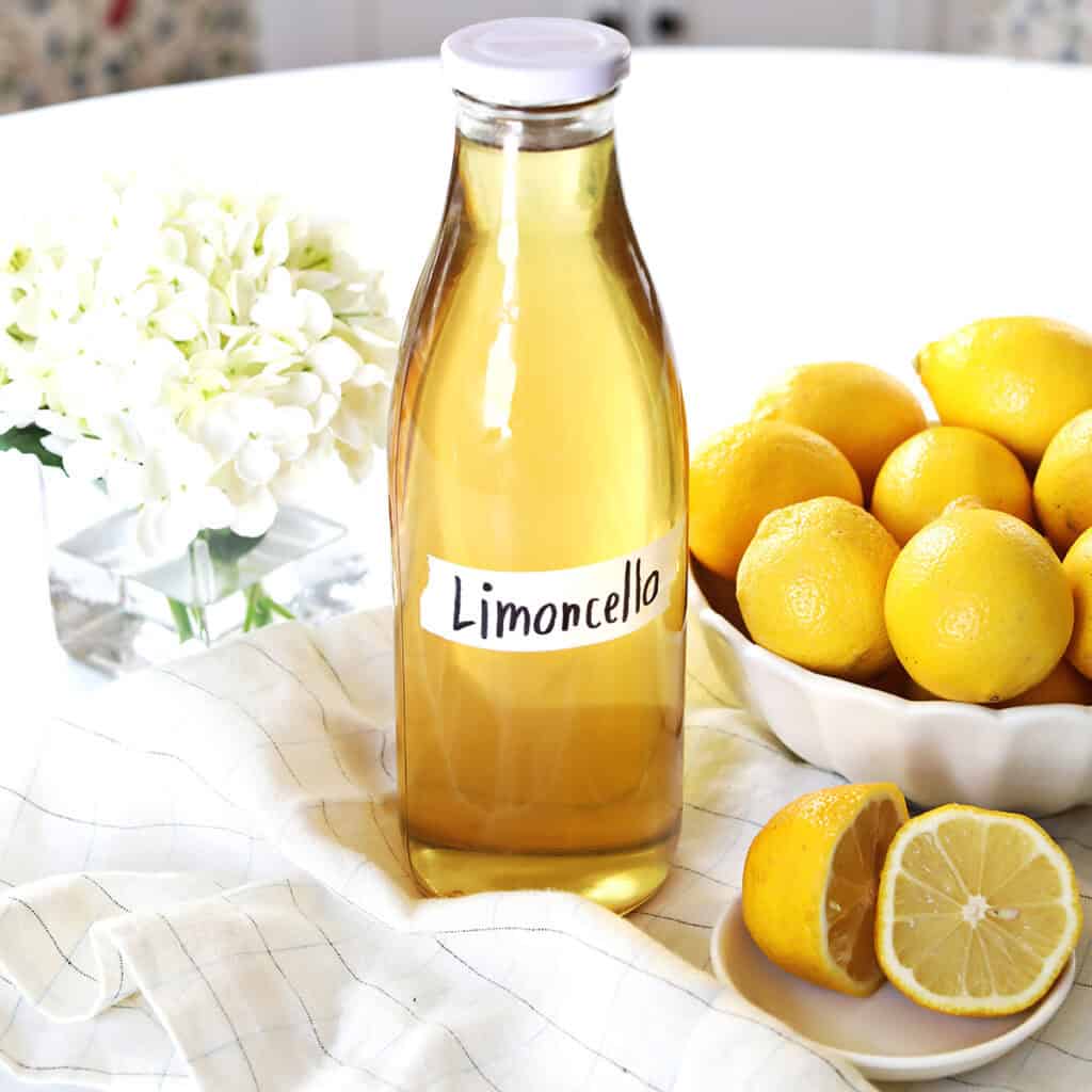 A bottle labeled "Limoncello" placed on a cloth next to a bowl of whole and sliced lemons, with a white flower arrangement in the background.