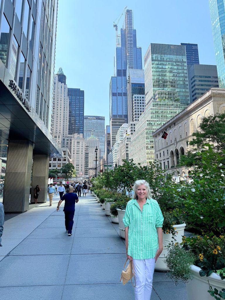 A person in a green striped shirt stands on a city sidewalk with tall buildings and greenery in the background on a sunny day.